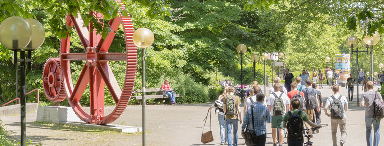 Studenten im Sommer auf dem Campus Nord, rote Zahnräder im Hintergrund